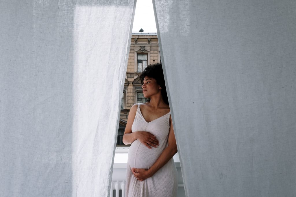 A Black expectant mother standing by a window and holding her baby bump
