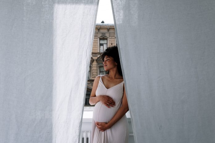 A Black expectant mother standing by a window and holding her baby bump A Black expectant mother standing by a window and holding her baby bump
