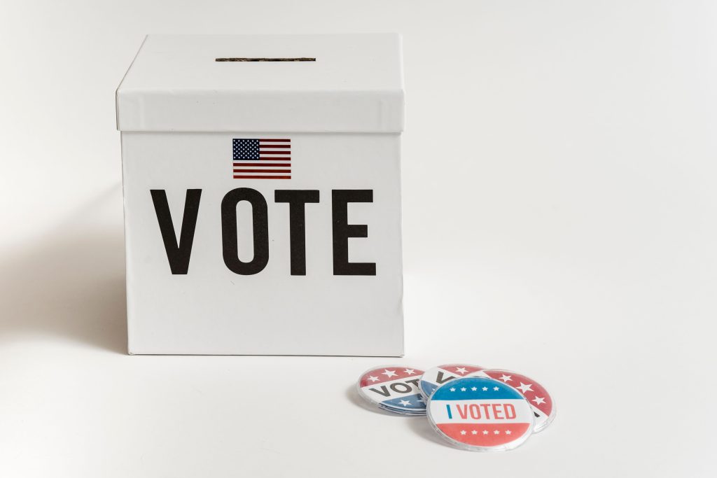 A white ballot box beside an American flag and I Voted badges.