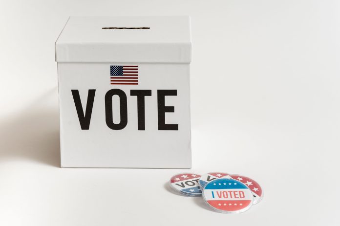 A white ballot box beside an American flag and I Voted badges.