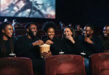 Black Movies in Theaters This Fall: Oct. and Nov. 2026 Dates to Save Black friends laughing and sharing popcorn in a dark movie theater lit by the screen