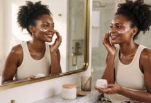 Fall Skin Care for Melanin-Rich Skin: A Dermatologist-Backed Routine A Black woman with deep brown skin applying moisturizer to her cheek in a bathroom mirror